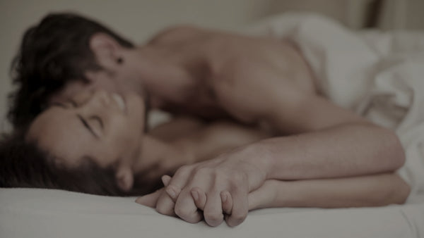 Man lying on top of a smiling woman in bed whispering his desires into her ear and holding her hand. 