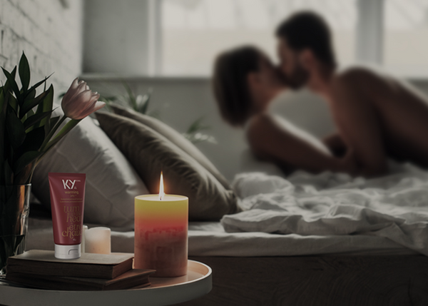 A couple in bed next to a lit candle beside a bottle of K-Y® Lubricant - Warming® Gel and plant.