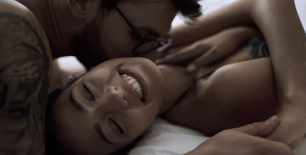 Un homme portant des lunettes embrasse le cou de sa partenaire souriante, dans le lit. 