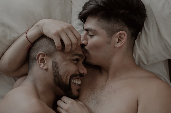 A couple laying on their backs in bed, embracing each other and smiling with a soft kiss.