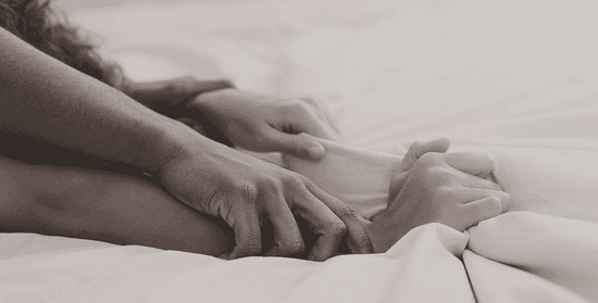 Neutral-toned close-up of a man holding a woman's wrists down on white bed sheets, demonstrating an intense intimate moment.