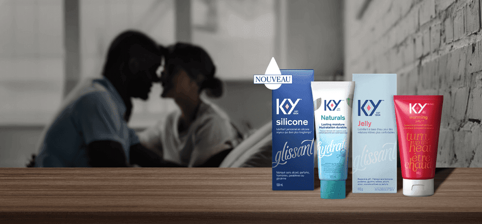 Des bouteilles de lubrifiant K-Y — Silicone, de lubrifiant K-Y — Hydratant+ gel intime Naturals et de lubrifiant K-Y — Gel Warming posées sur une tablette en bois située devant un couple assis face à face sur un lit.