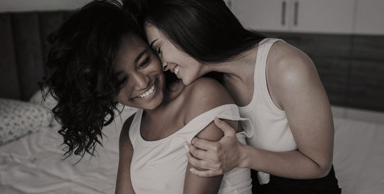 A loving embrace between two women in a softly lit bedroom, both beaming with happiness.