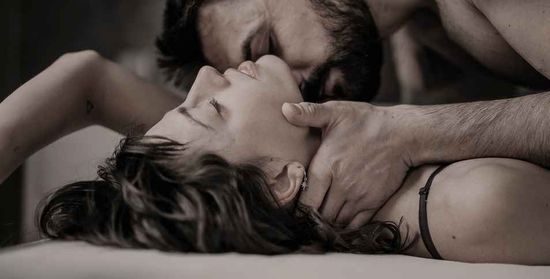 A couple in bed share a passionate kiss, the man’s hand resting on the woman’s neck as he kisses her intimately.