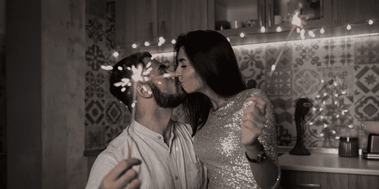 Un couple s’embrassant en tenant des cierges magiques dans une cuisine chaleureuse, décorée de guirlandes lumineuses.