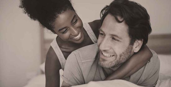 Neutral-toned photo of a woman hugging a man from behind while they relax together in a cozy bedroom