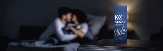 Gros plan d’une boîte du lubrifiant K-Y — Silicone devant un couple s’enlaçant dans un lit.